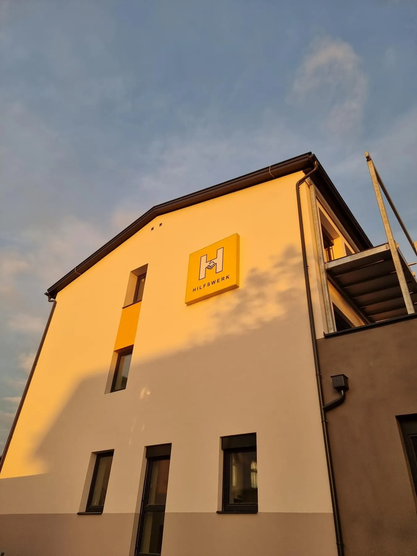 Gebäudeansicht mit Hilfswerk-Leuchtschild im Abendlicht