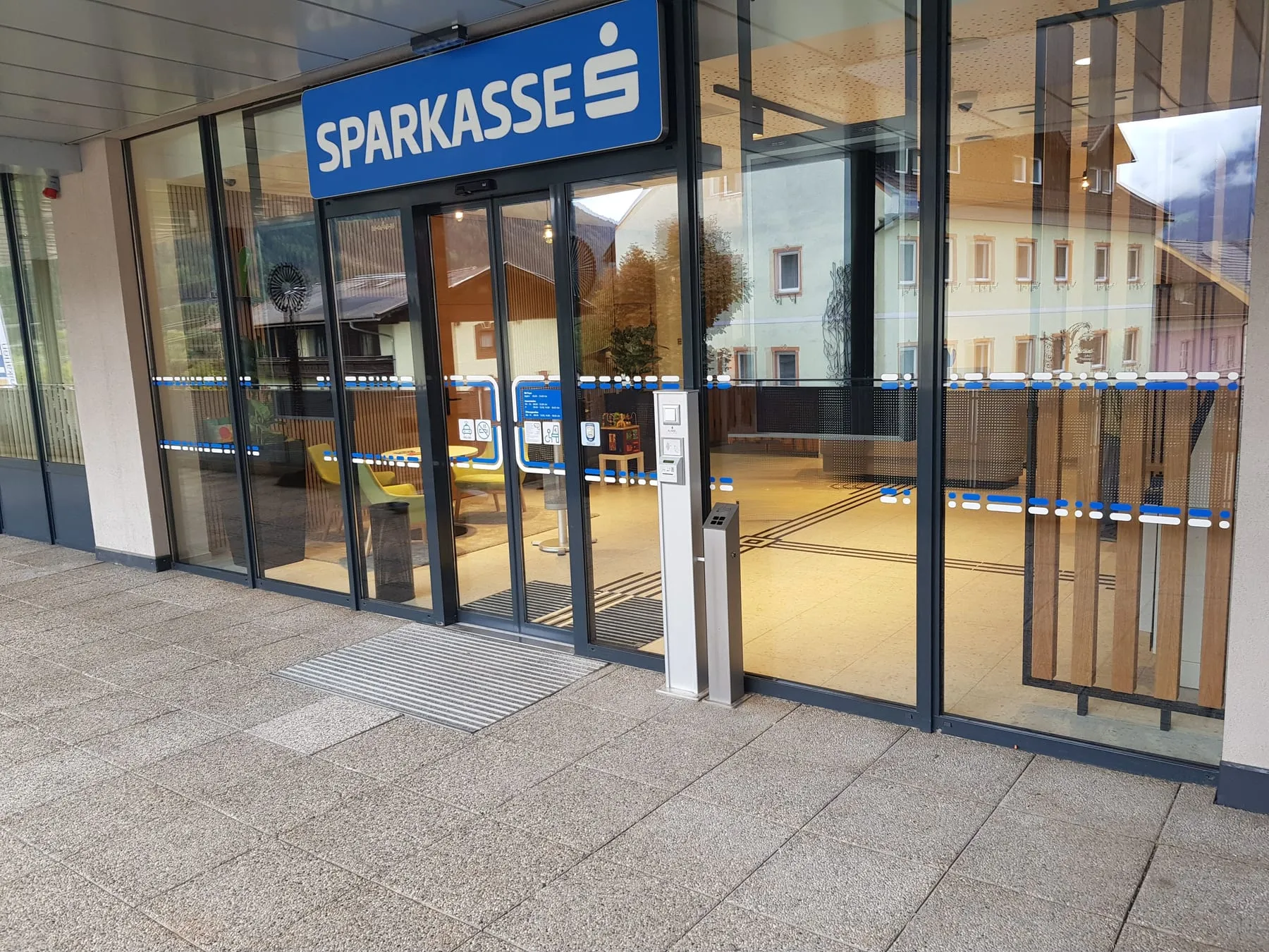 Eingang einer Sparkasse-Filiale: Schild über der Tür, blau-weiße Markierung an der Glastür, drinnen Leitstreifen am Boden