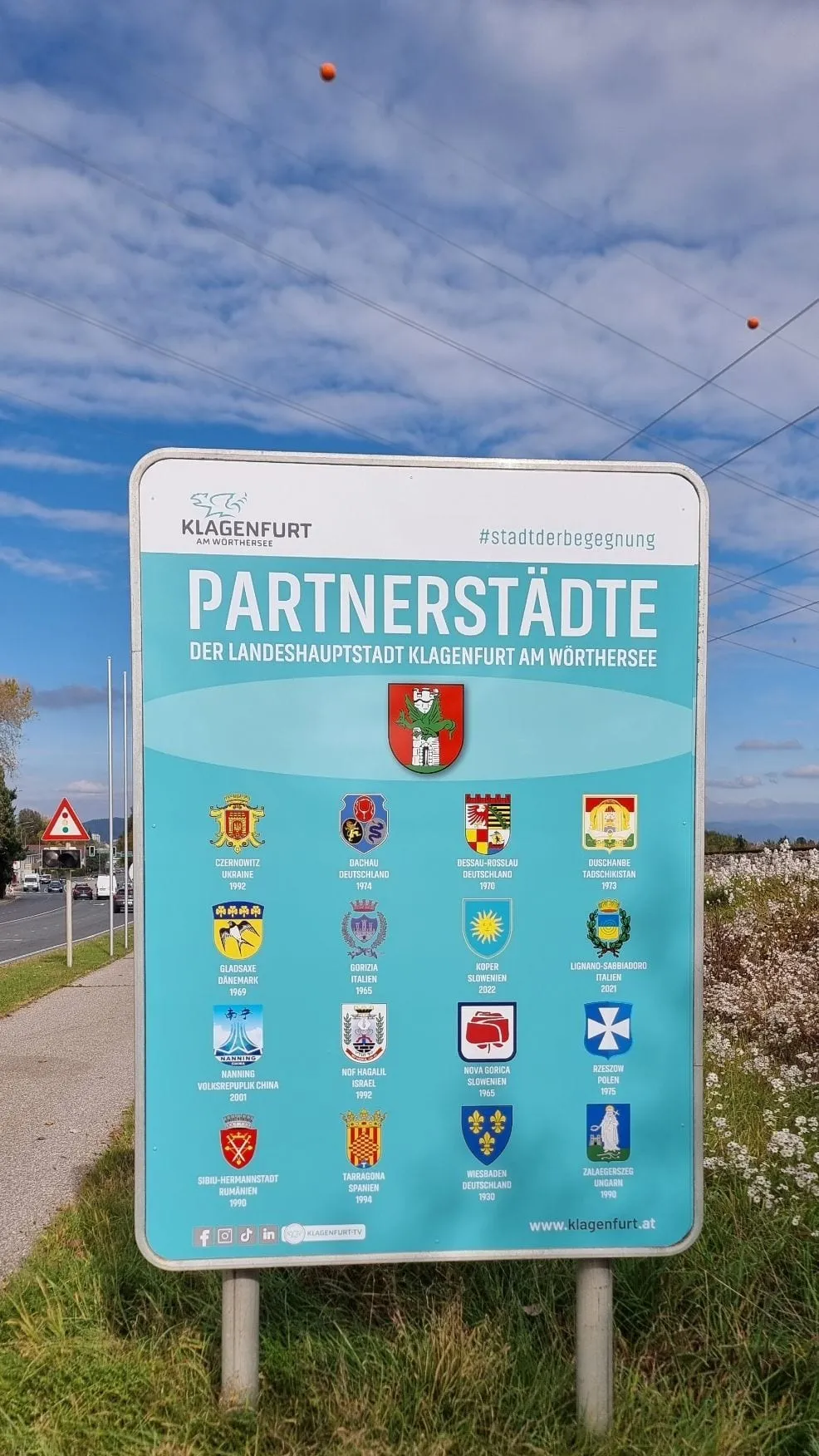 Tafel „Partnerstädte der Landeshauptstadt Klagenfurt am Wörthersee“ mit Wappen, auf zwei Stehern vor blauem Himmel