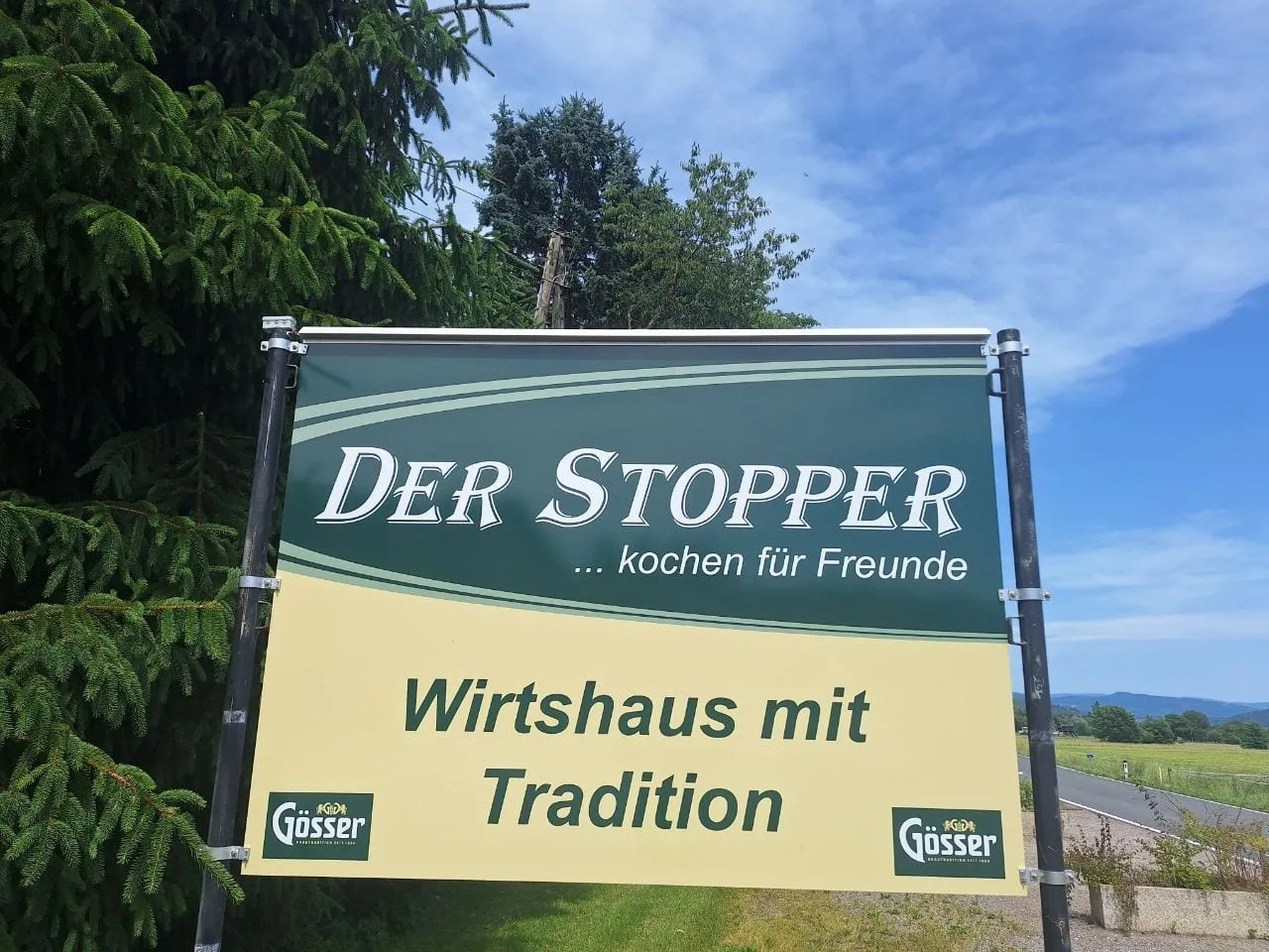 Werbetafel eines Wirtshauses mit Gösser-Logos an zwei Pfosten vor blauem Himmel