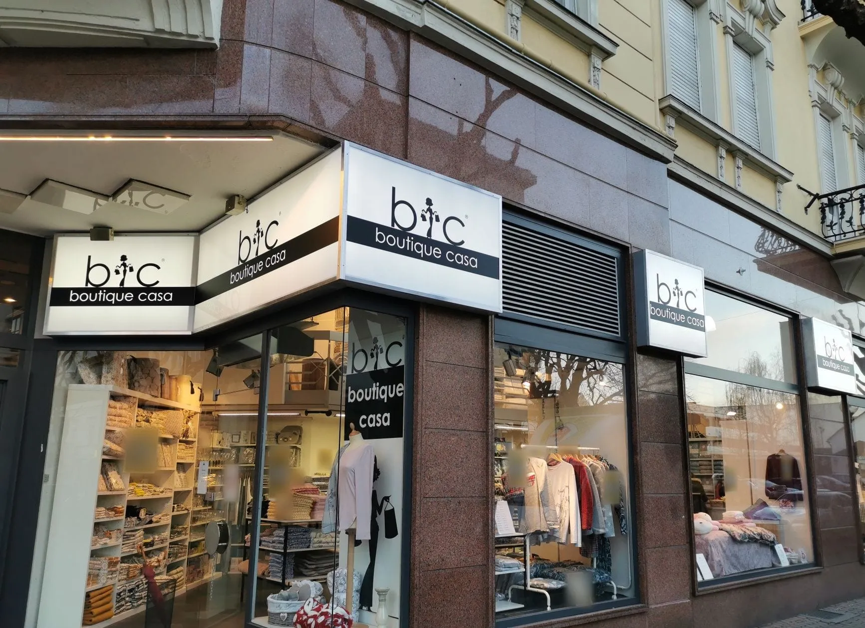 Beleuchtete Leuchtkästen von b+c boutique casa an einer Eckfassade in der Dämmerung
