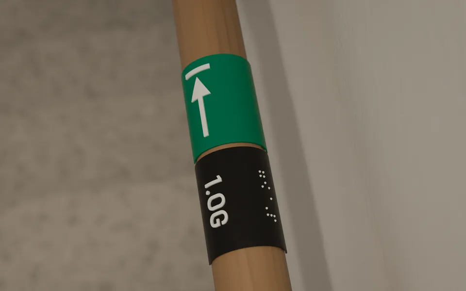 Beispielansicht: Nahaufnahme eines Handlaufschilds. Ein grünes Feld mit Pfeil zum Ausgang und ein schwarzes Feld mit „1.OG“ in erhabener Schrift, daneben zur Wand hin Brailleschrift.