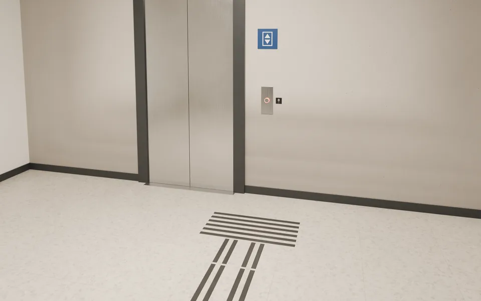 Beispielansicht: Ein Leitstreifen ohne Mittelsteg führt zu einem Feld aus quer liegenden Stegen vor dem Lift, mittig unter der Ruftaste. Daneben die Geschoßzahl zum Ertasten.